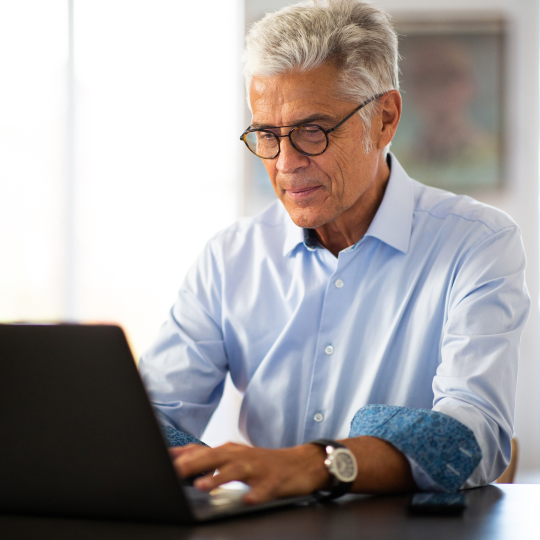 A smiling older man with grey hair and glasses works intently on a laptop, wearing a light blue shirt.